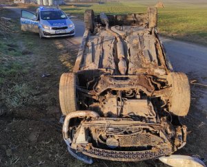 Wywrócony na dach samochód marki peugeot 206 w tle stoi policyjny radiowóz